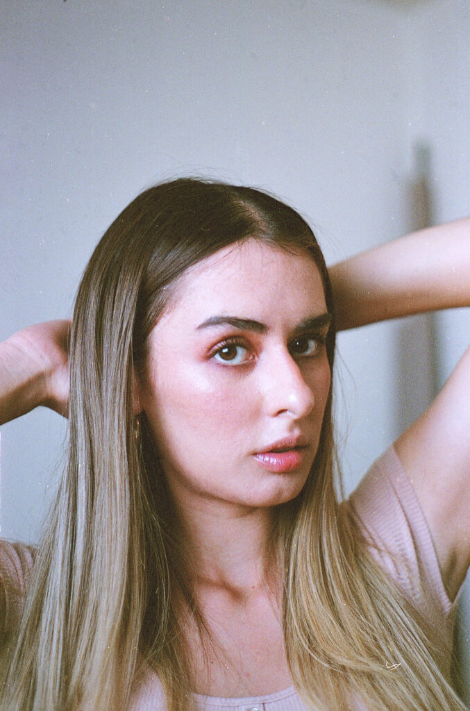 Film Photo of Marintia Bustos a model with long brown hair wearing a light pink top posing with her hands behind her head against a neutral background.